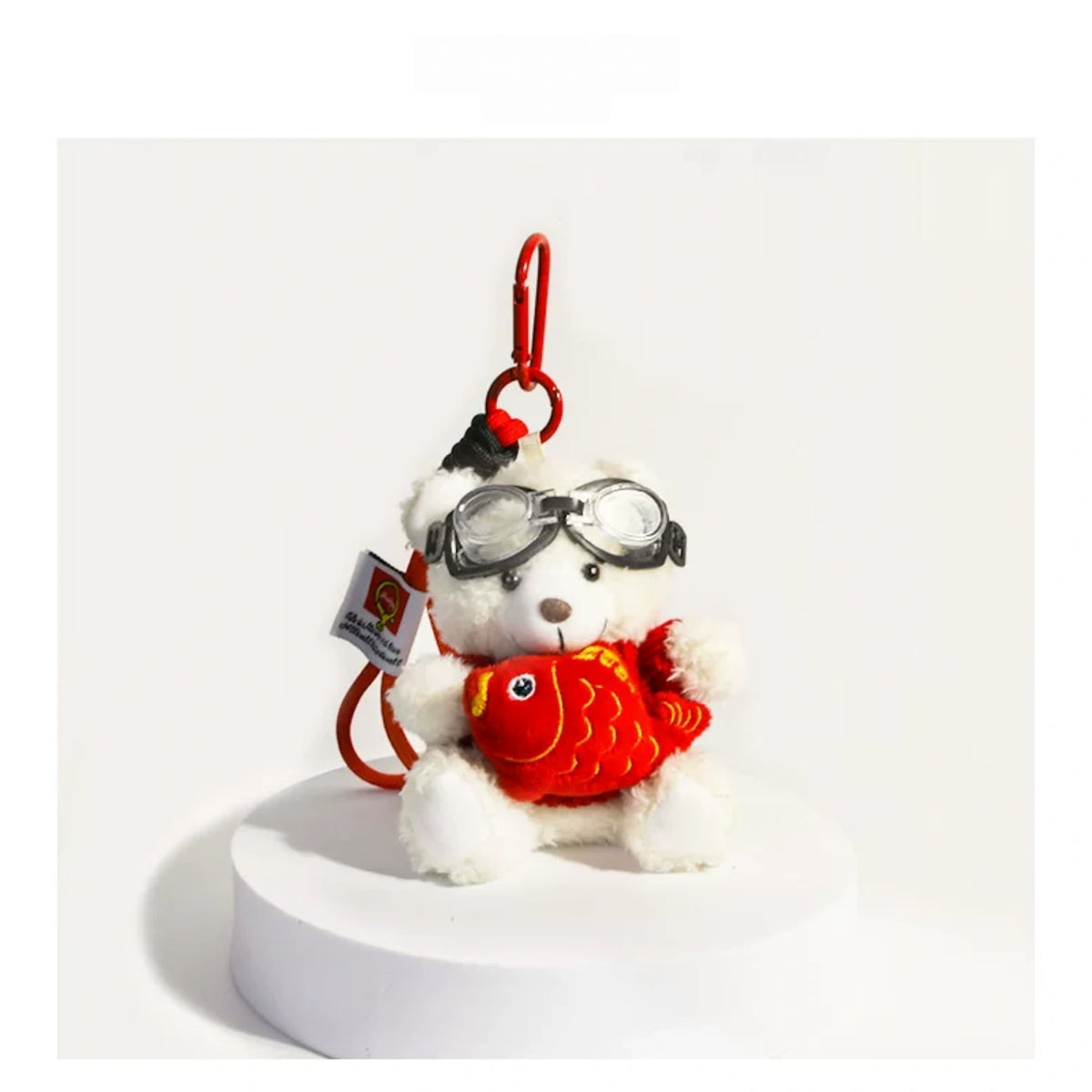 White plush Spirit Bear keychain wearing goggles, holding a red fish charm, on a white pedestal