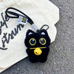 Black plush Yoyo keychain with a yellow bell collar, shown on a canvas tote bag outdoors