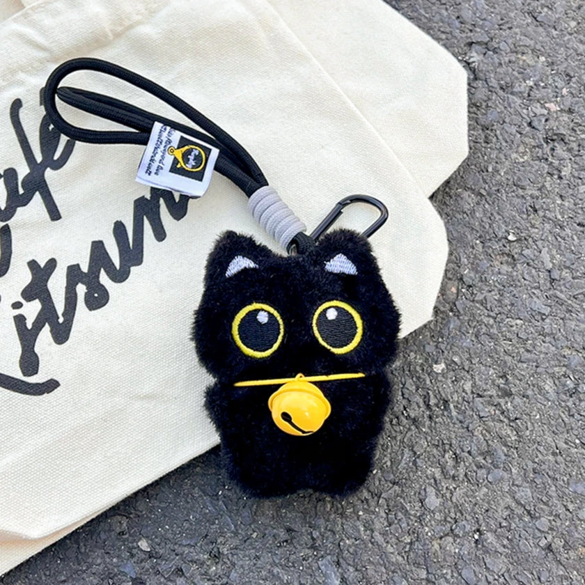 Black plush Yoyo keychain with a yellow bell collar, shown on a canvas tote bag outdoors