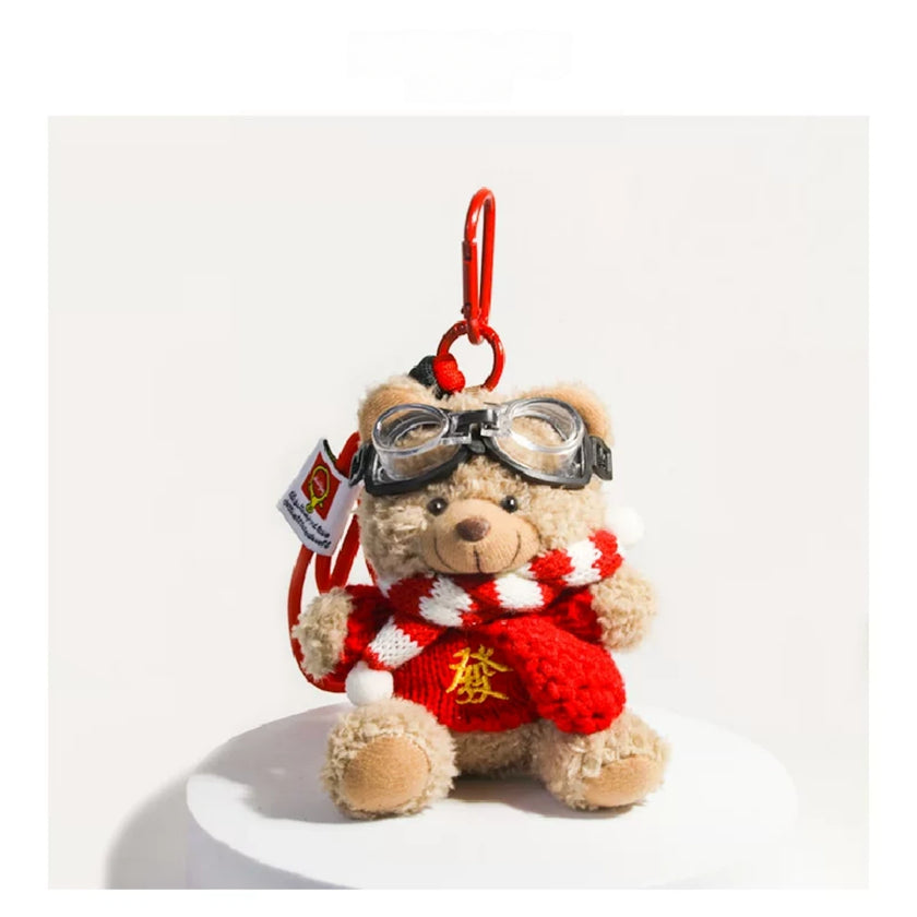 Tan plush Spirit Bear keychain wearing goggles and a red sweater with gold characters