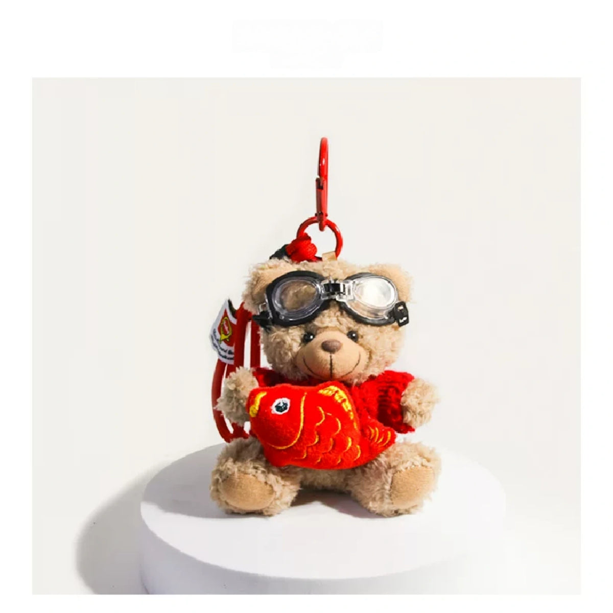 Tan plush Spirit Bear keychain wearing goggles, holding a red fish charm
