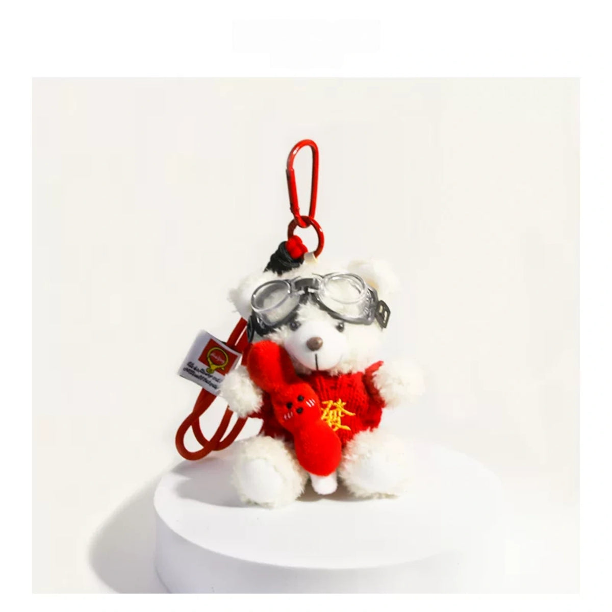 White plush Spirit Bear keychain wearing goggles and holding a red charm, on a white pedestal