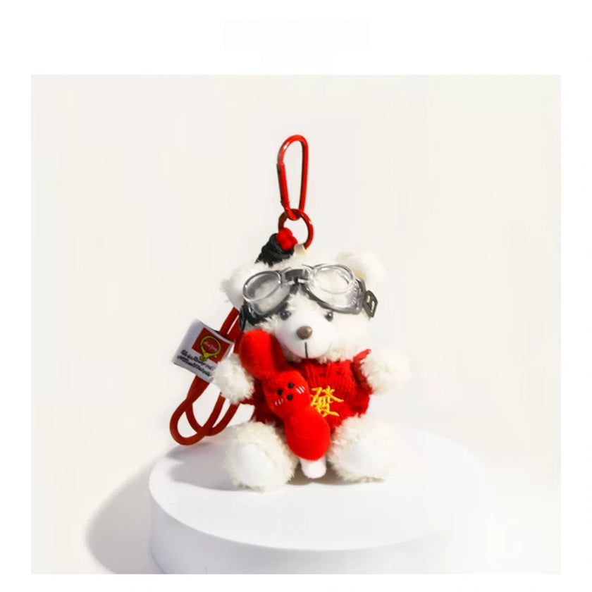 White plush Spirit Bear keychain wearing goggles and holding a red charm, on a white pedestal