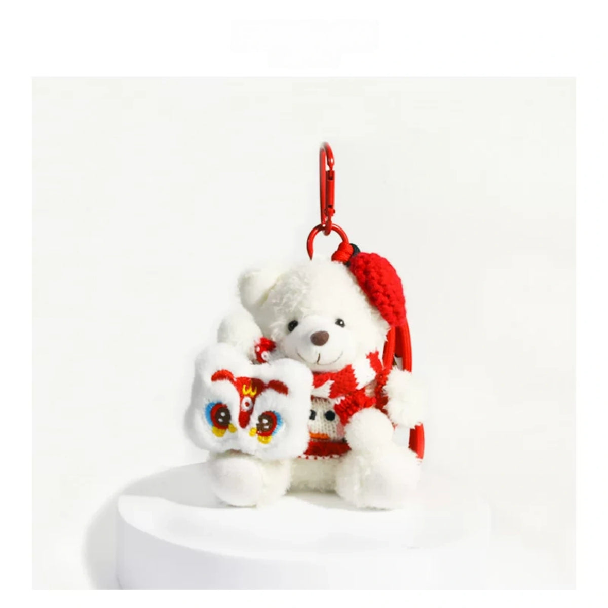 White plush Spirit Bear keychain wearing a red beret, holding a Lucky Lion charm, on a white pedestal
