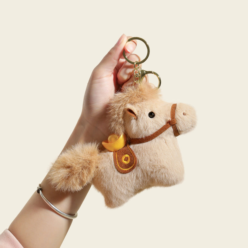 Hand holding a tan plush Heavenly Steed horse keychain with a gold ingot charm